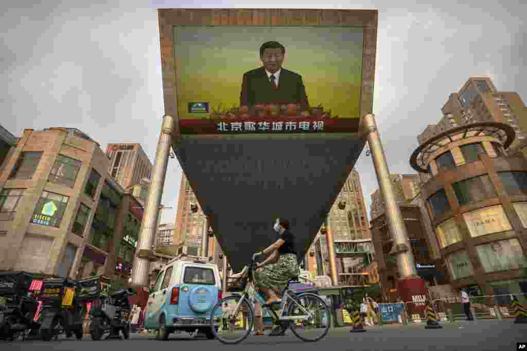 A woman wearing a face mask rides a bicycle past a large television screen at a Beijing shopping center displaying Chinese state television news coverage of Chinese President Xi Jinping's visit to Hong Kong.
