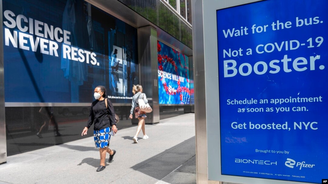 FILE - Pedestrians walk past a signs hanging outside Pfizer headquarters in New York and one hanging at a bus stop encouraging the COVID-19 booster, May 23, 2022. 